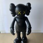 A black KAWS Companion figure with yellow 'X' eyes stands against a white and cardboard background.