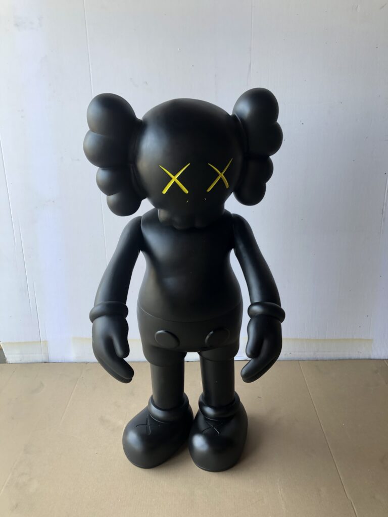 A black KAWS Companion figure with yellow 'X' eyes stands against a white and cardboard background.