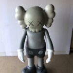 A large, grey KAWS Companion figure with X's for eyes and white gloves, standing on a brown surface.