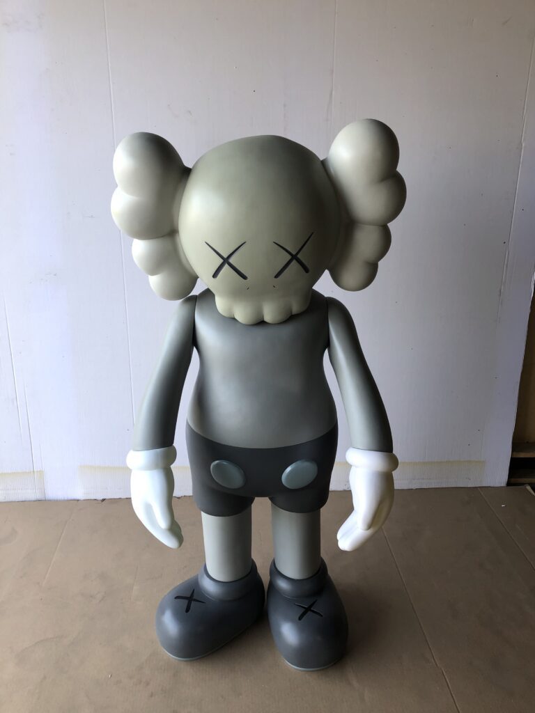 A large, grey KAWS Companion figure with X's for eyes and white gloves, standing on a brown surface.