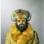 A man with a beard, yellow sunglasses, and a fur hat and coat looks directly at the viewer.