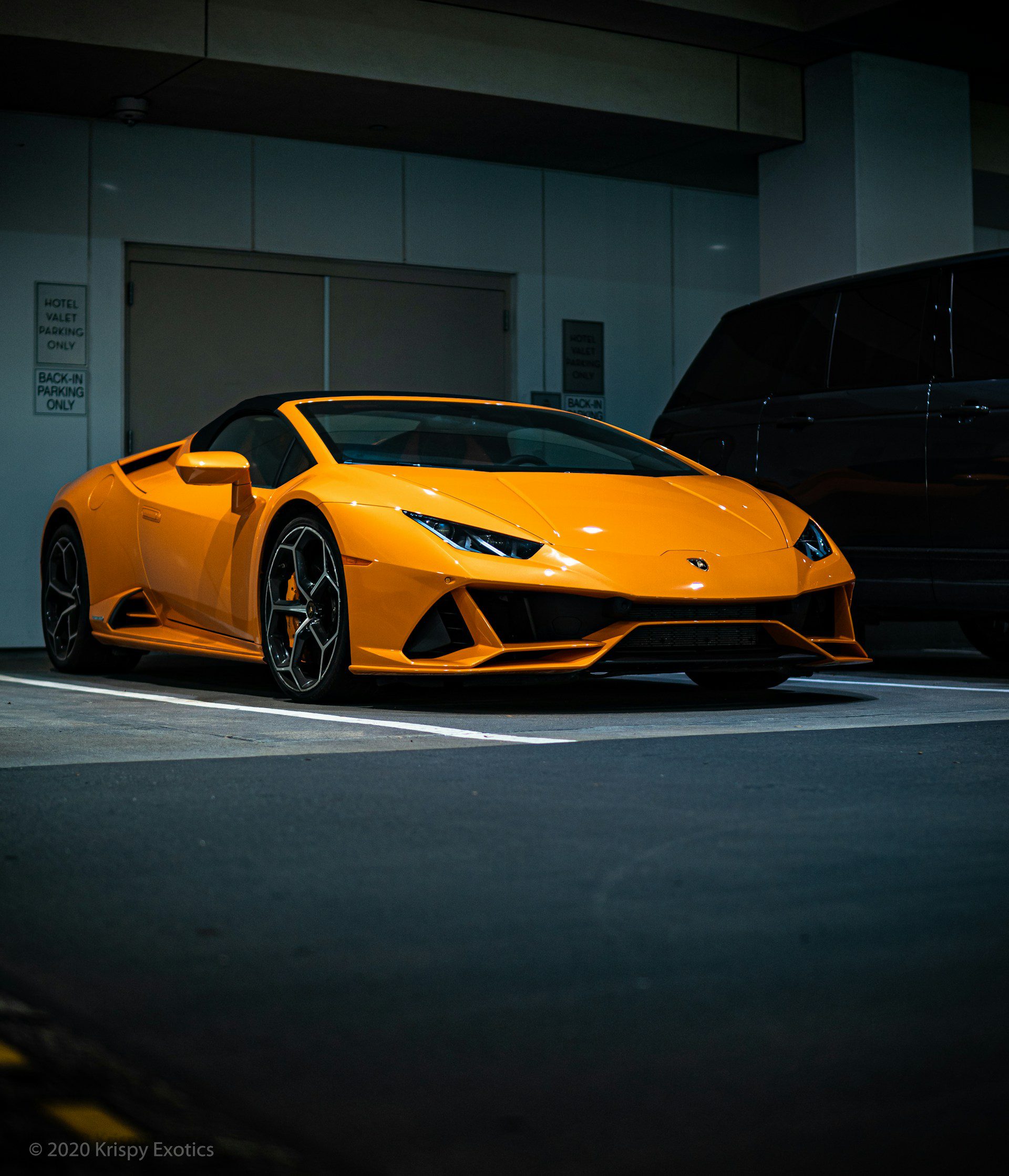 A bright orange Lamborghini Huracan Evo Spyder is parked in an underground garage next to a black SUV.