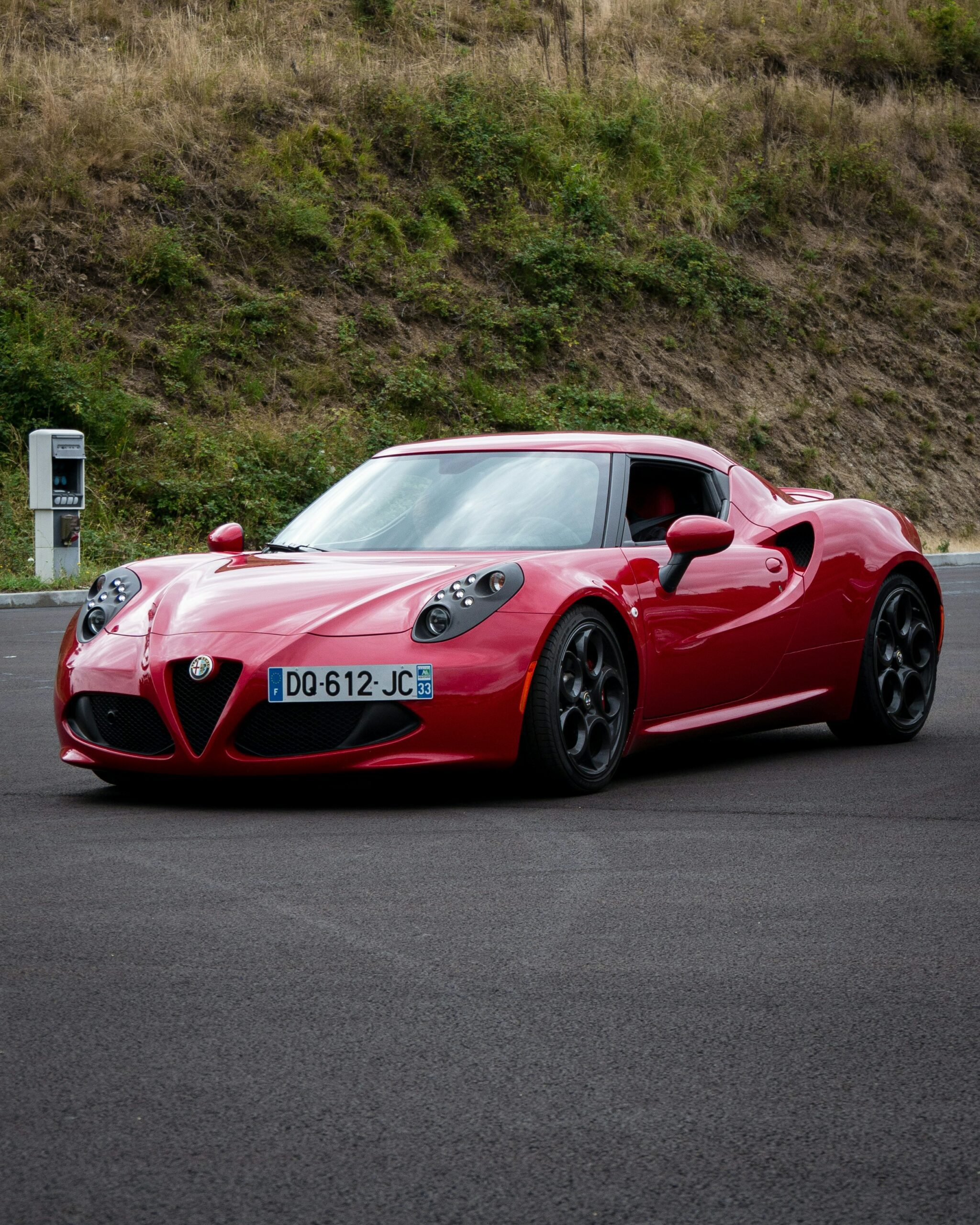 A sleek red Alfa Romeo 4C sports car parked on asphalt with a grassy hill and charging station in the background.