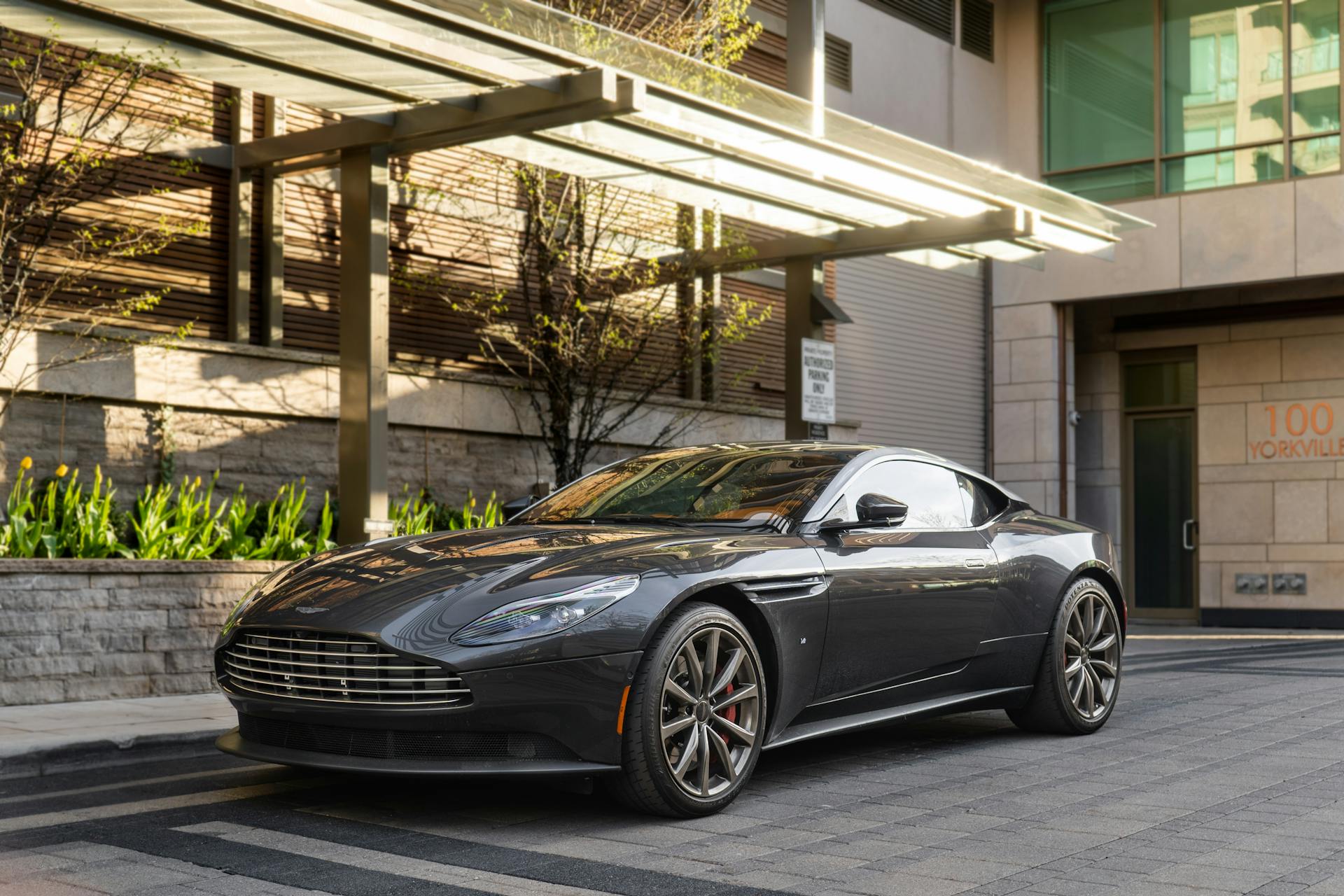 A sleek, dark gray Aston Martin DB11 parked on a paved street in front of a modern building with trees.