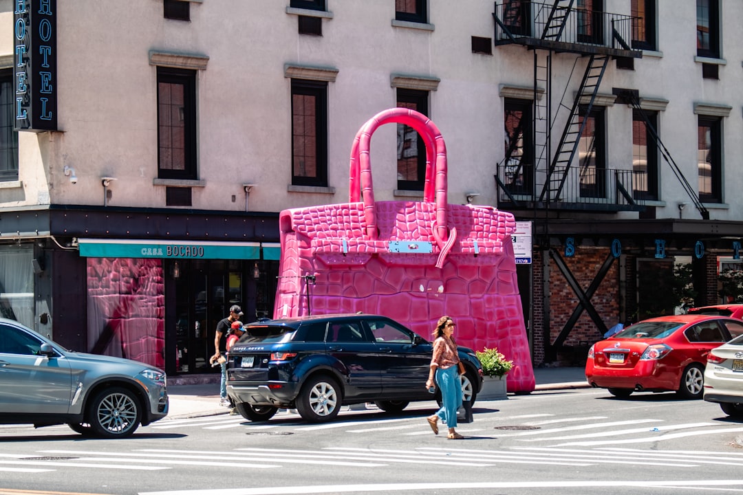 Birkin-bag-better-asset-than-car - borro Oversized pink handbag sculpture on a city street sidewalk, with pedestrians crossing and cars parked along the curb.