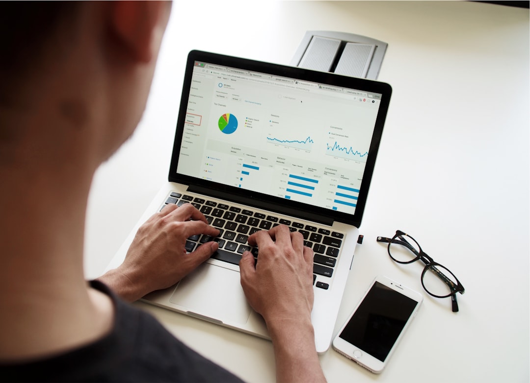 Person typing on a laptop showing a data analytics dashboard with charts, on a white desk with a phone and glasses nearby