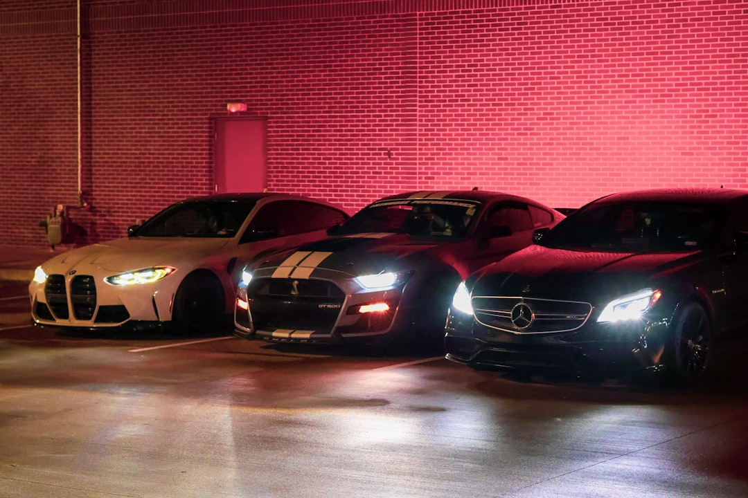 Three sports cars parked side by side at night against a red-lit brick wall: white bmw on the left, black shelby gt500 with white stripes in the middle, and black mercedes on the right.