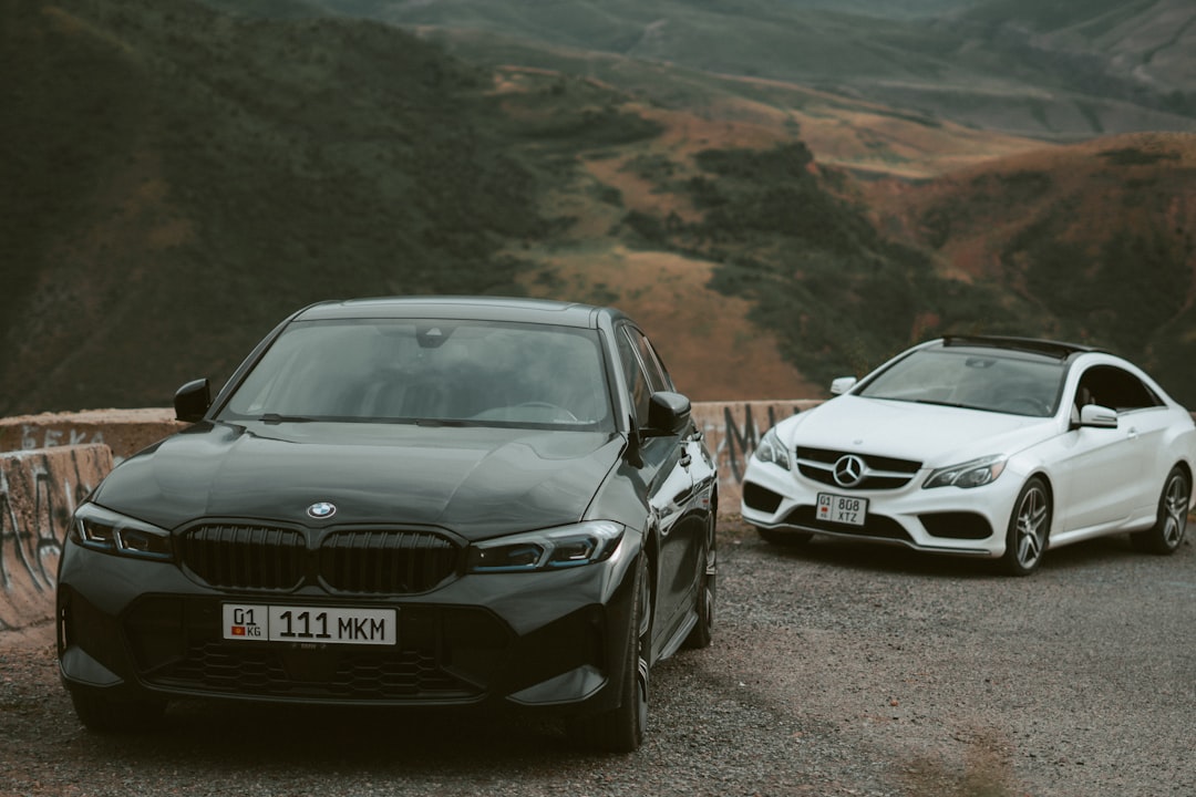 Two luxury cars, a black bmw in the foreground and a white mercedes behind, parked on a gravel mountain road with hills in the background. ”] }{