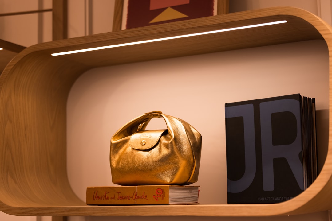 Gold metallic handbag displayed on a small stack of books inside a curved wooden shelf with warm lighting behind it.
