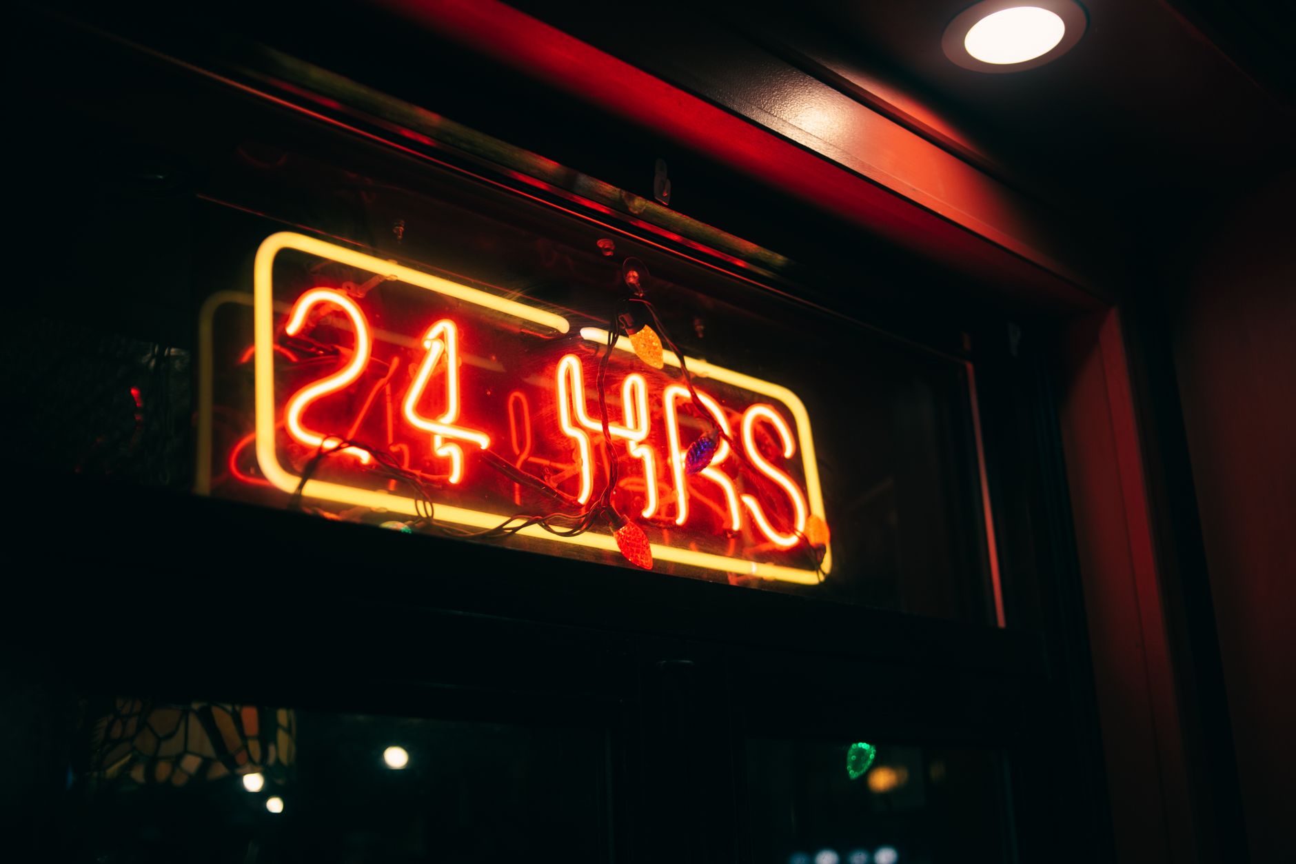 Neon sign in a storefront window glowing red and yellow, reading '24 hrs' at night, suggesting the shop is open 24 hours.