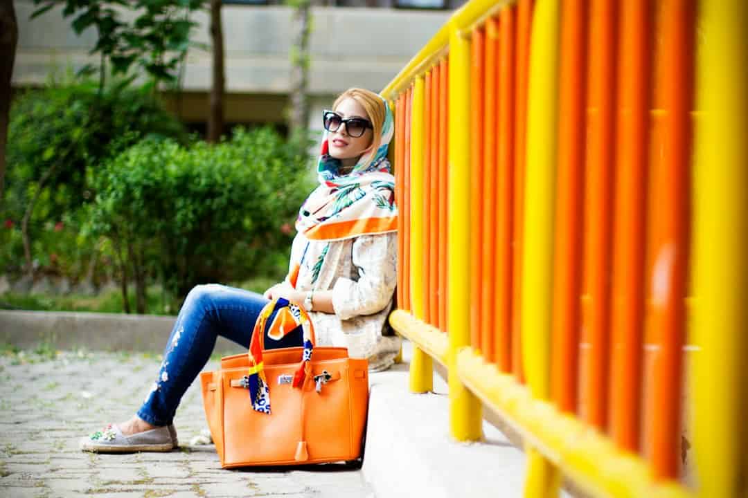 Fashionable woman sitting by vibrant yellow and orange fence with orange handbag.