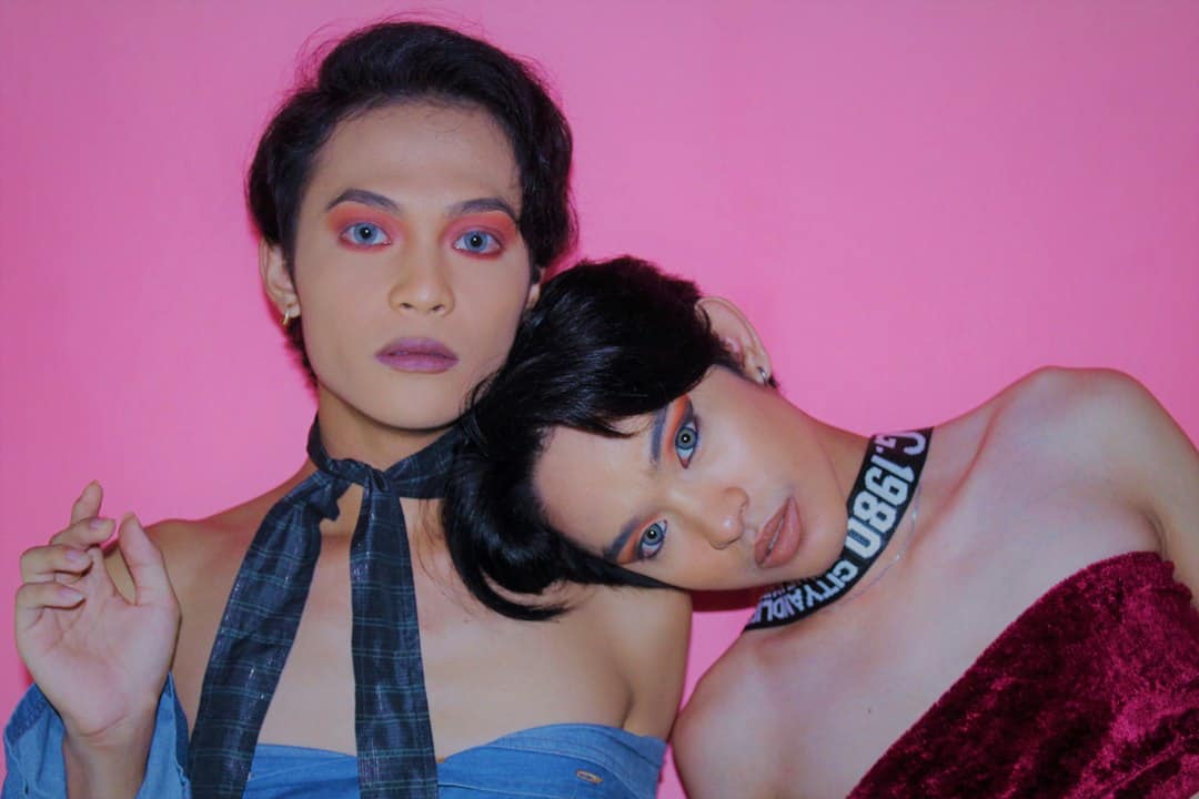 Two women with bold makeup and stylish outfits against pink background.